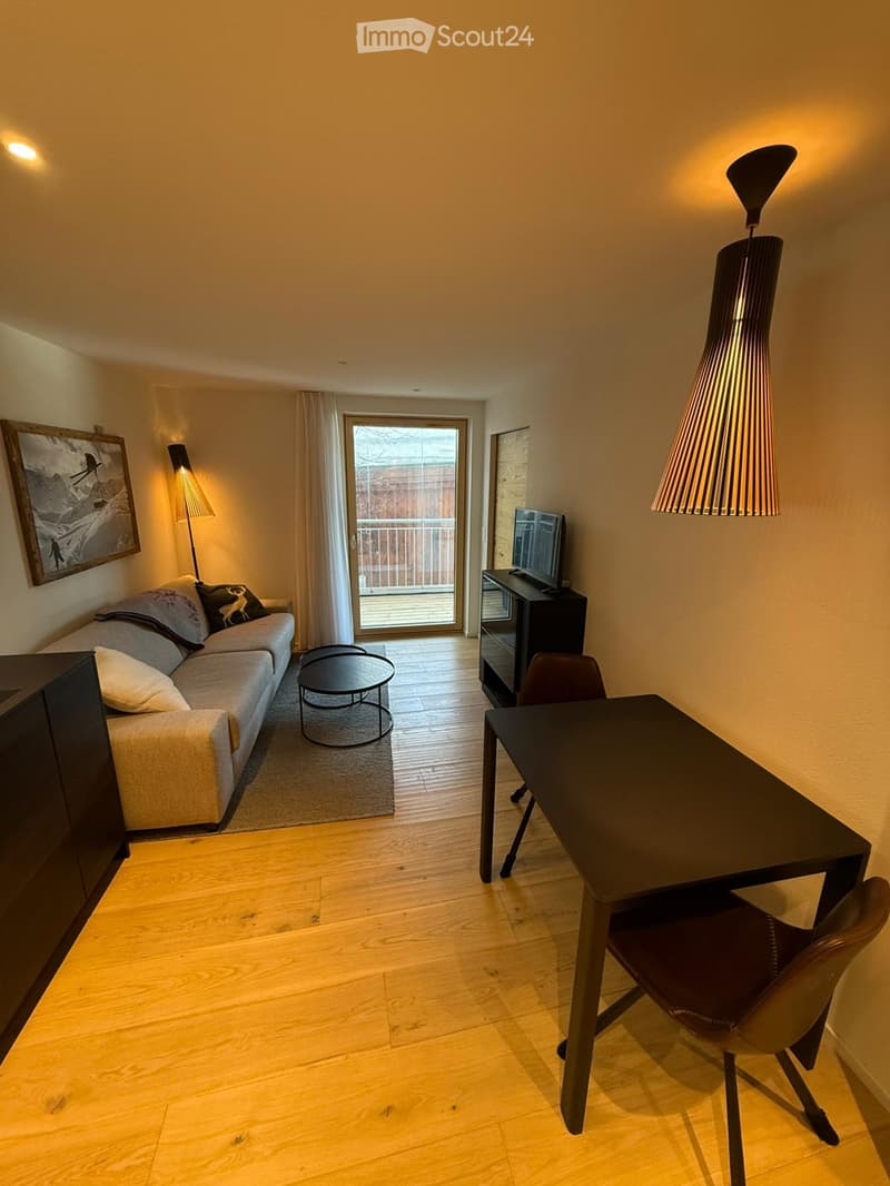 A modern living room with a sofa, coffee table, TV, and a desk area with a chair and table. The room features wooden flooring, warm lighting, and a large window with a view of a balcony.