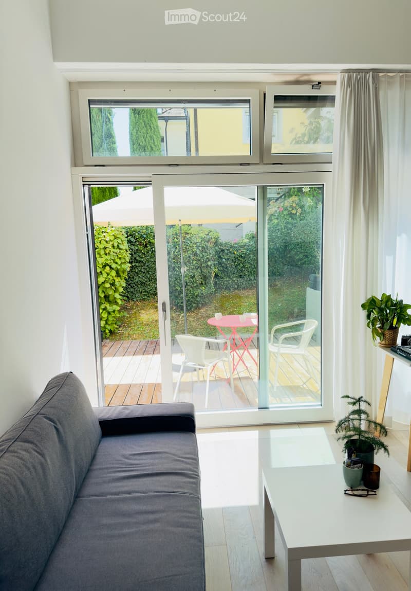 Modern living room with a grey sofa, white coffee table, and large windows leading to a balcony with outdoor furniture.