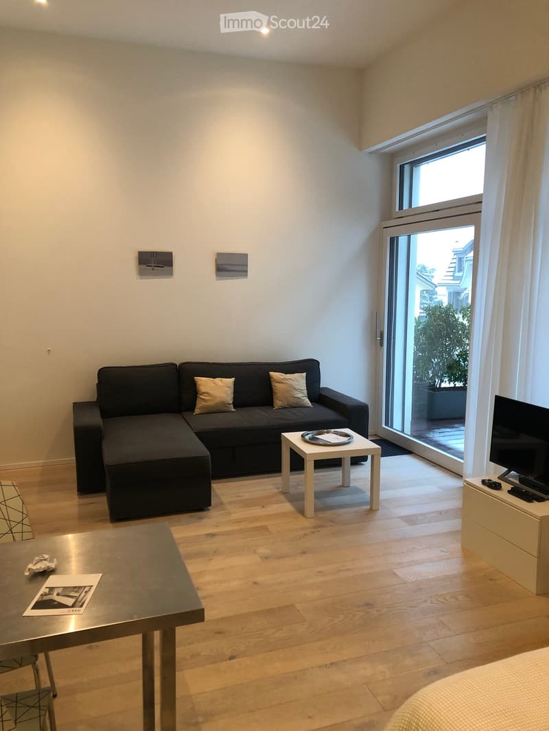 Modern living room with light wooden flooring, a dark grey sectional sofa with two throw pillows, a small white coffee table, a larger rectangular table with a magazine and crumpled paper, and a TV on a stand near a large window with white curtains.