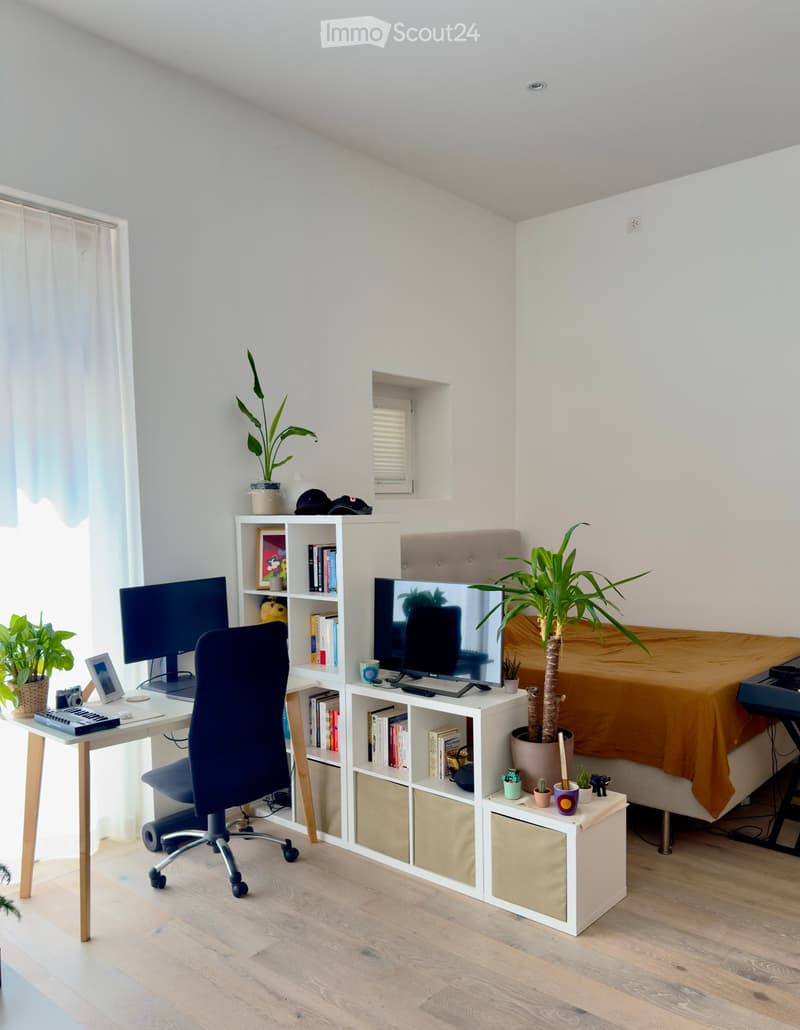 A modern, well-lit room with a desk setup, a bed, and a shelf filled with books and decorative items. The room features a large window with a curtain, a potted plant, and a piano in the corner.