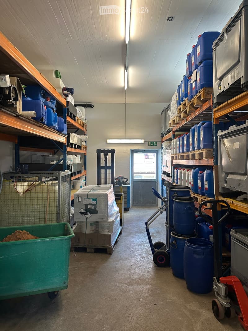 A storage area with shelves holding blue barrels, a hand truck, a cart, and a green container with material inside. The space has a concrete floor and a door leading outside.
