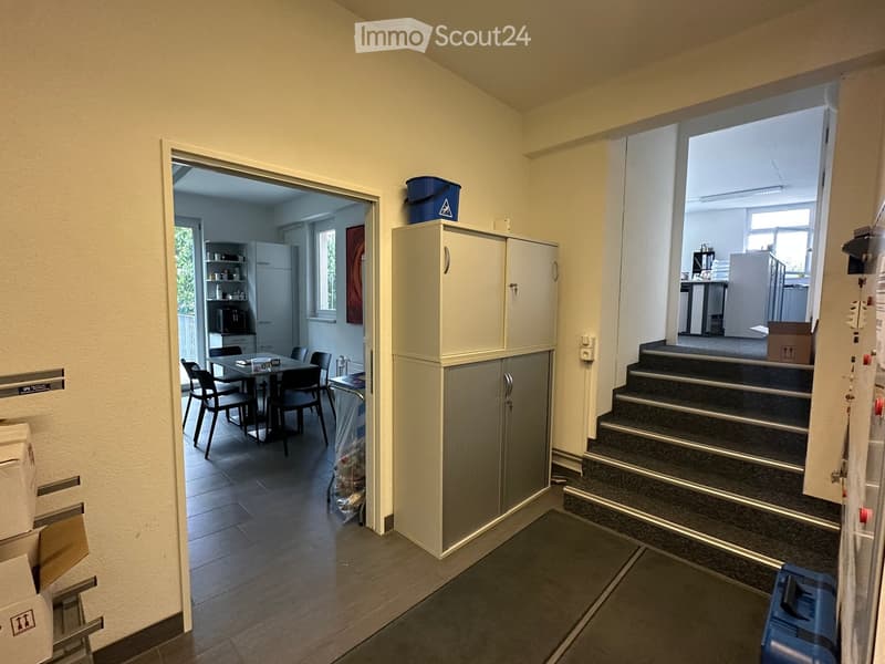 Hallway with tiled floor, white walls, and a staircase leading to another level. Visible are a tall cabinet, a small table with chairs, and a doorway to a kitchen area.
