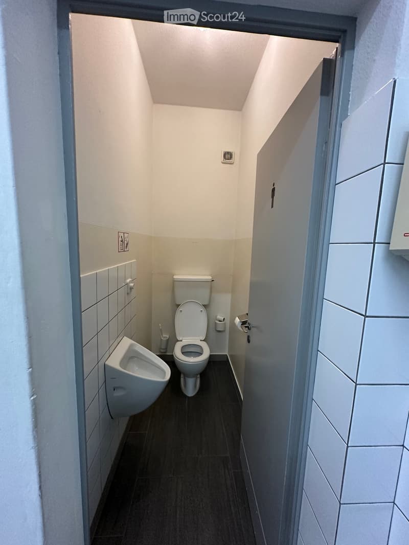 Modern bathroom with a toilet, urinal, and toilet paper holder. The walls are tiled, and the floor is dark.