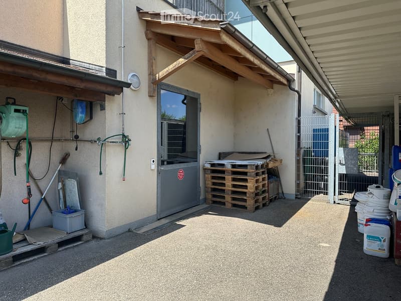Outdoor area with a covered entrance, stacked pallets, storage containers, and a hose reel mounted on the wall.