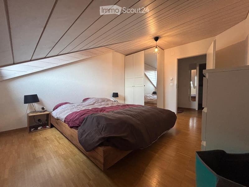 Bright, modern bedroom with wooden flooring, a large bed with colorful bedding, a nightstand with a lamp, and a white wardrobe. The room features skylights and warm lighting.