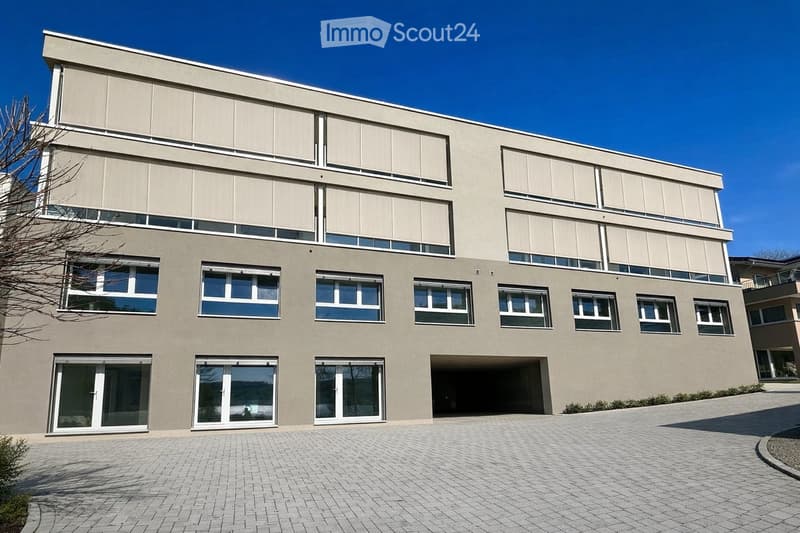 Modern two-story building with large windows, a flat roof, and a paved driveway. Features include a garage entrance and multiple floor-to-ceiling windows.