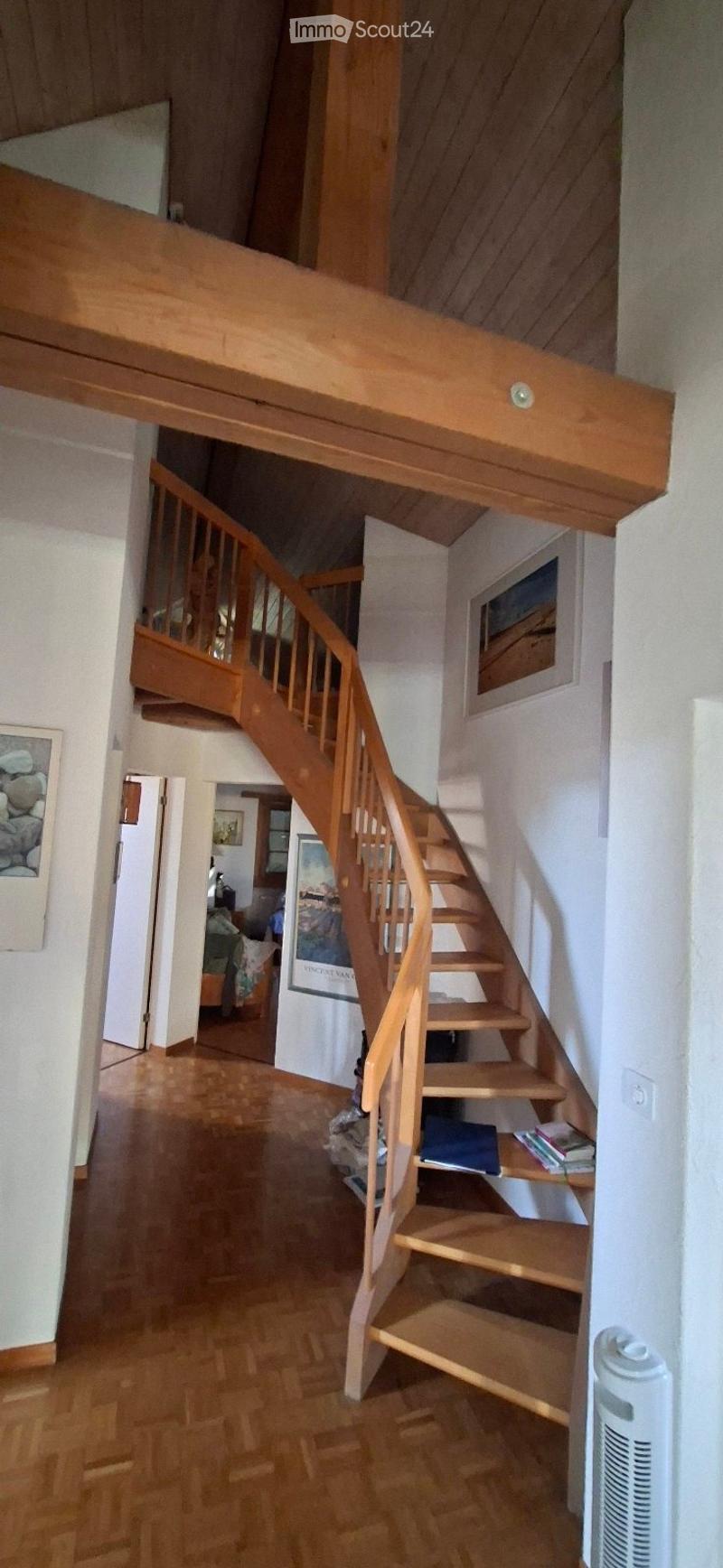 Wooden staircase leading to an upper level, with wooden railings and steps, light-colored walls, and a small shelf with books and a blue bag on the landing.