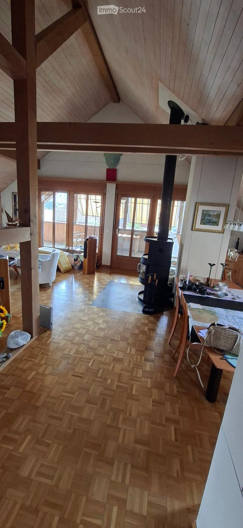 Spacious living room with wooden herringbone flooring, white walls, and exposed wooden beams. Features a wood stove, sofa, chair, and a small table with items on it.