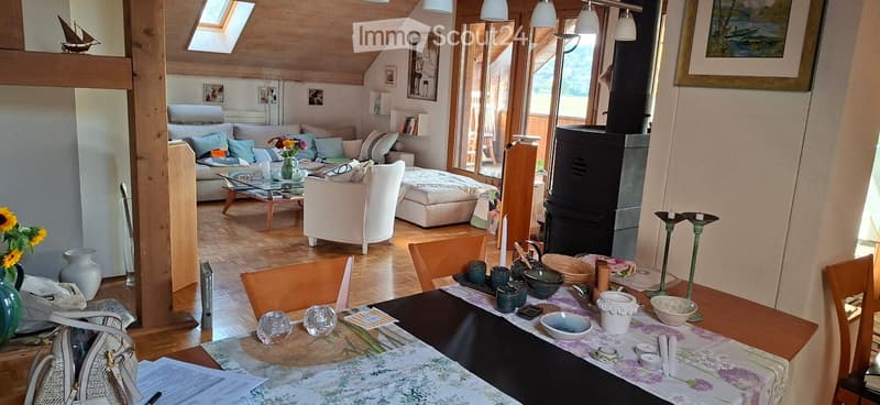 Bright, open living and dining area with wooden flooring, large windows, a sofa, coffee table, armchair, dining table with chairs, and decorative items including vases and candles.