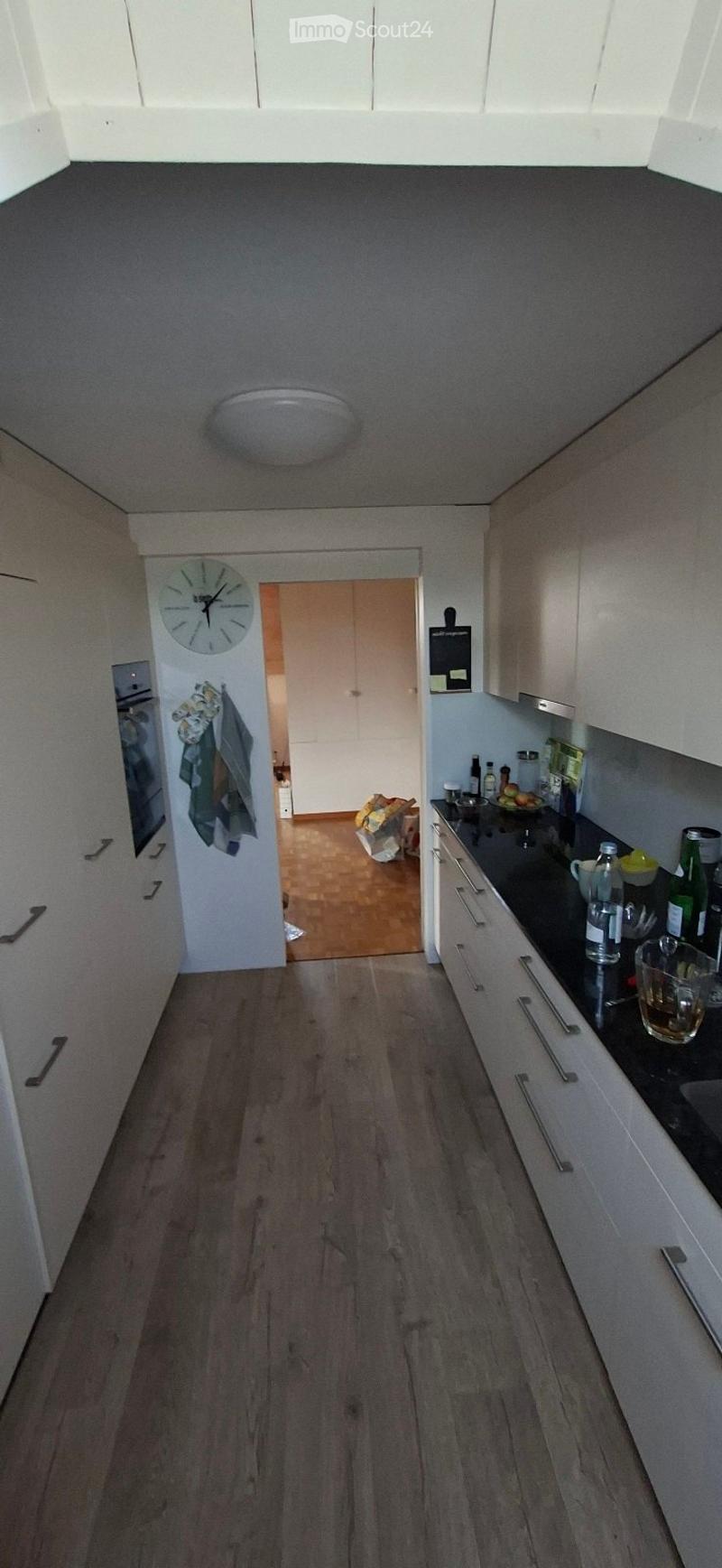 Modern kitchen with white cabinets, dark countertop, and wooden flooring. Visible items include a clock, bottles, cups, and a bowl on the counter.