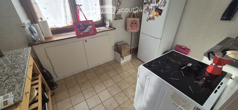 A small kitchen with white cabinets, a black induction stove, a refrigerator, and a granite countertop. There are various items on the windowsill and a handbag hanging on the wall.