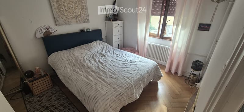 Cozy bedroom with wooden flooring, a large bed with a dark blue headboard, a nightstand with a lamp, a mirror, a decorative fan, and a wicker basket near the window.