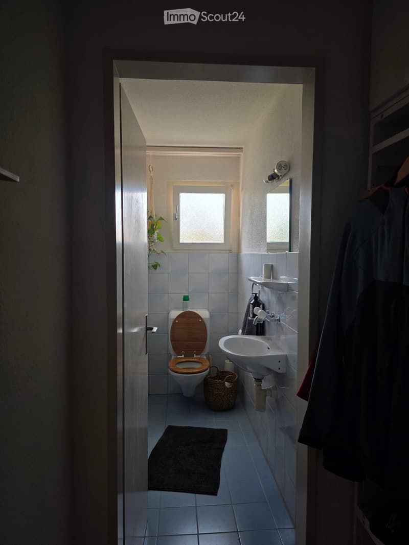 Modern bathroom with a toilet, sink, mirror, and wicker basket. The space features tiled walls and floor, a window for natural light, and a dark-colored rug in front of the toilet.