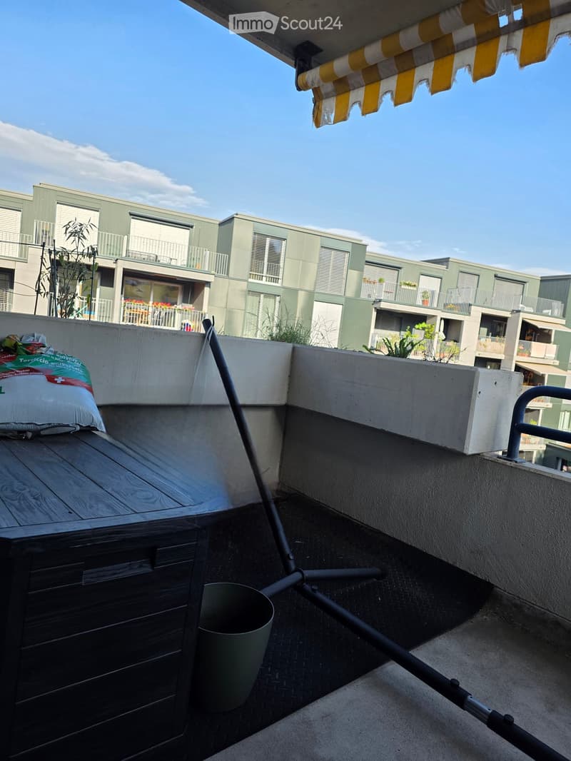 Balcony with wooden table, metal railing, and awning, overlooking a modern apartment complex with balconies and greenery.