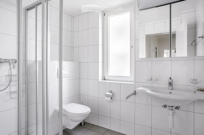 Modern bathroom with white tiled walls and floor, featuring a toilet, sink, mirror, and a glass-enclosed shower area.