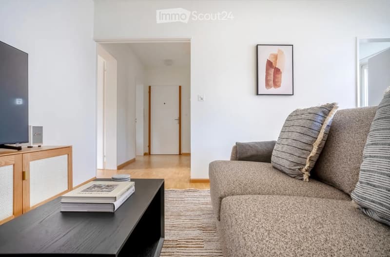 Modern living room with a grey sofa, coffee table with books, TV stand, and a hallway leading to another room.