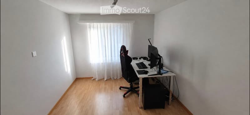 A small, well-lit room with wooden flooring, a desk with a computer setup, and a chair.