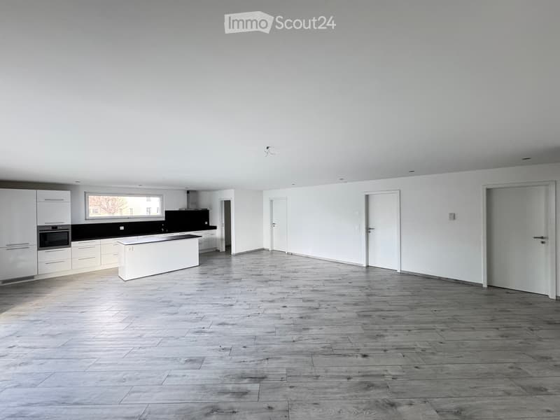 Open-plan space with a modern kitchen area featuring white cabinets, a central island, and a black countertop. The main area has a wooden floor and white walls, with several doors leading to other rooms.