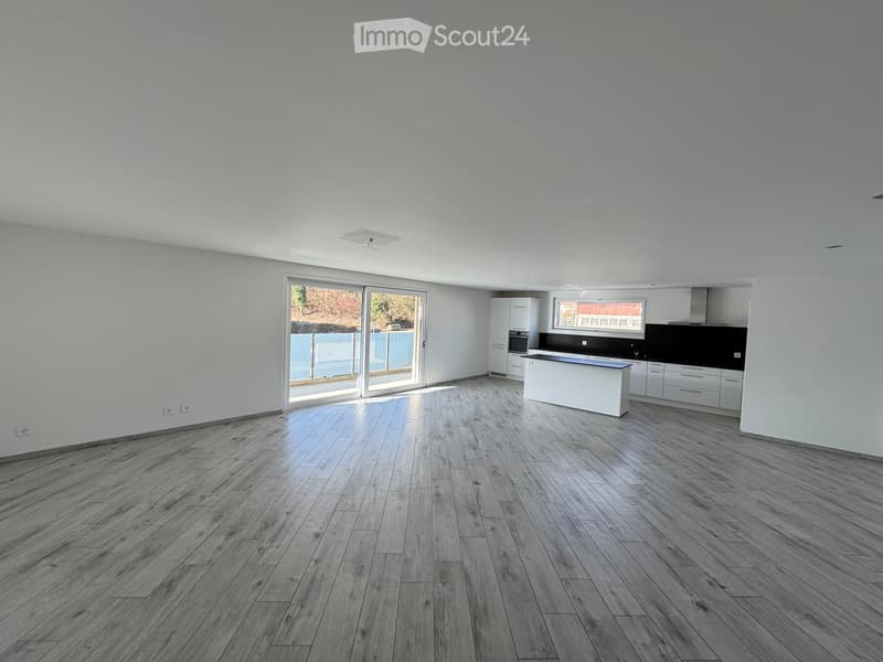 Open-plan living and dining area with wooden flooring, white walls, and large sliding glass doors leading to a balcony. Kitchen area with white cabinets and a black countertop is visible in the background.