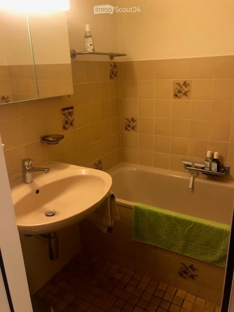 Bathroom with a sink, mirror, bathtub, and a green towel draped over the edge. A small shelf holds a bottle and other items.