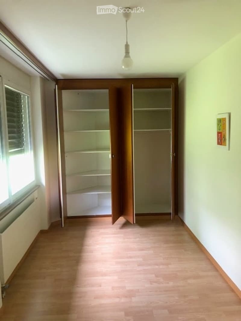 Empty room with wooden flooring, large window with blinds, built-in closet with open doors, and a single light fixture hanging from the ceiling.