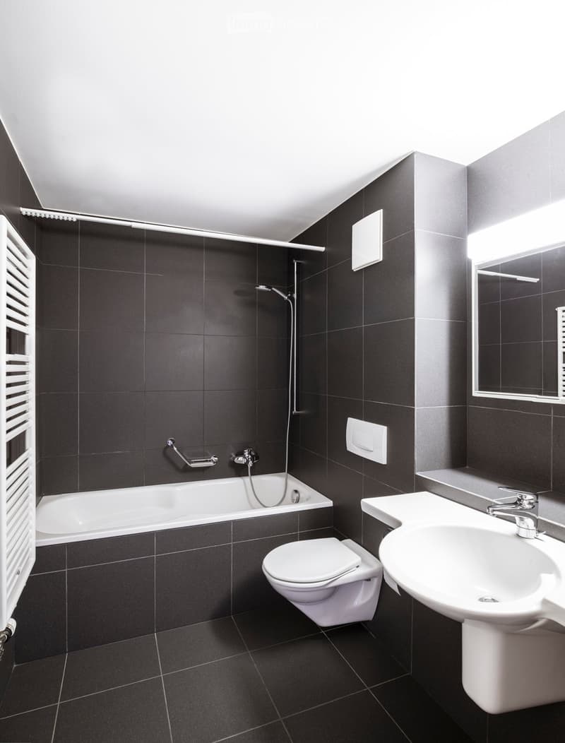 Modern bathroom with dark tiled walls and floor, featuring a bathtub, toilet, sink, and a mirror above the sink.
