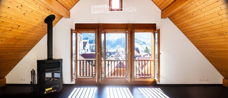 Bright, wooden-paneled room with a wood stove, large windows offering a scenic mountain view, and a balcony accessible through the windows.