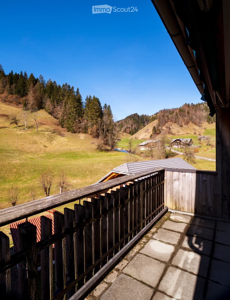 Balcony with wooden railing overlooking a scenic countryside with green fields, scattered trees, and distant houses.