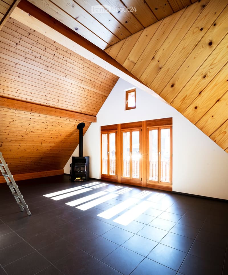 An attic space with wooden slanted ceilings, dark tiled floor, and large wooden-framed windows. A black wood-burning stove is positioned against the wall, and a ladder is visible on the left side.