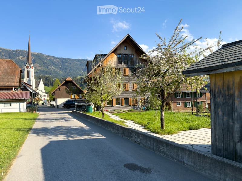 Village street with traditional wooden houses, a church tower, and surrounding mountains under a clear blue sky.