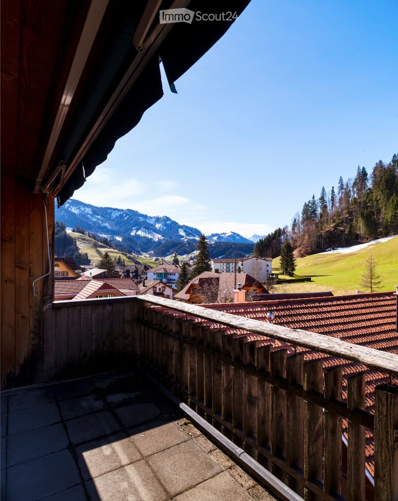 Balcony with wooden railing overlooking a village with houses, green hills, and snow-capped mountains in the background.