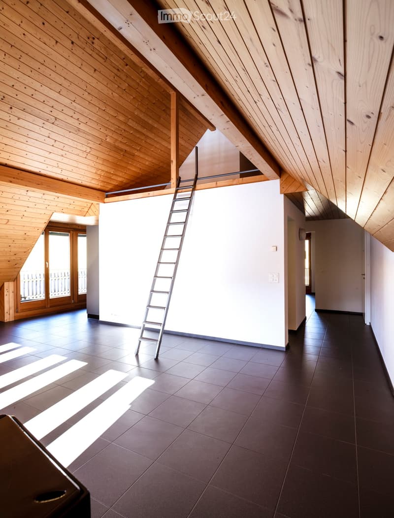 An empty attic space with wooden slanted ceilings, dark tiled floor, and a ladder leading to the upper level. Natural light streams in from a large window on the left side.