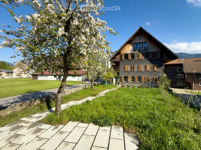 Traditional wooden house with multiple windows and shutters, surrounded by a well-maintained lawn and a blooming tree. The house features a pitched roof and is situated in a serene, green environment with a clear blue sky in the background.