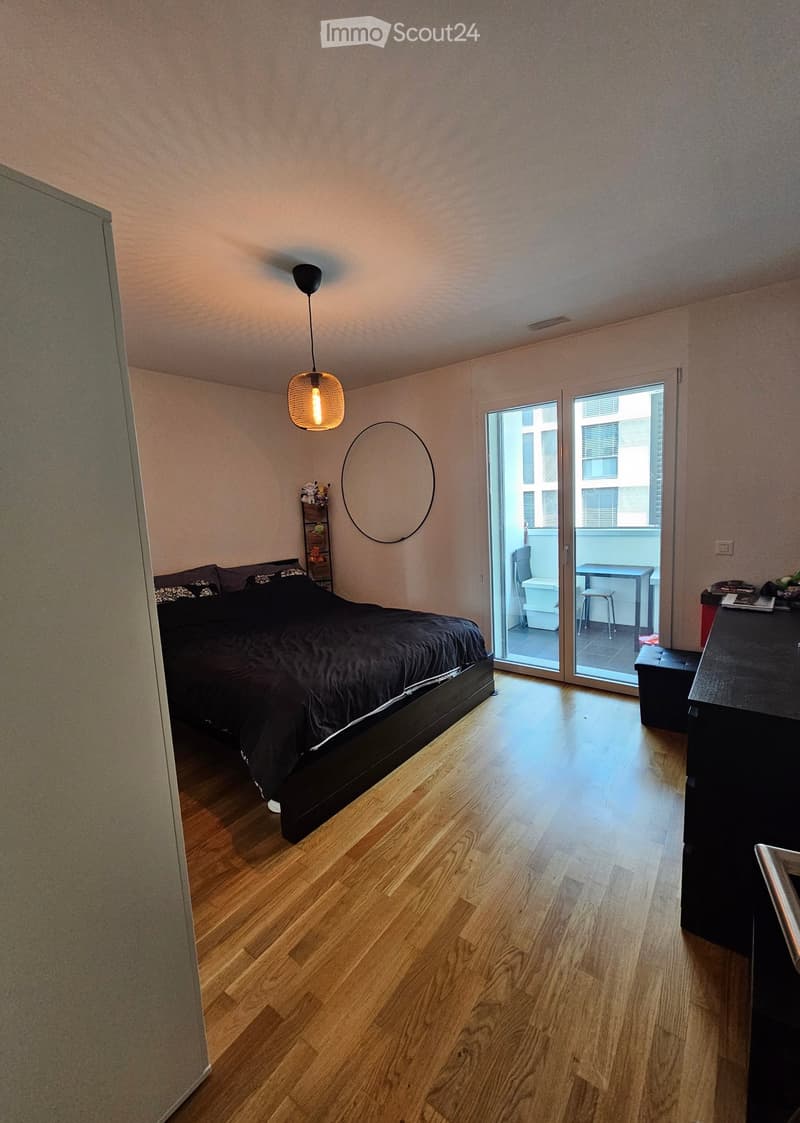 Modern bedroom with wooden flooring, a large bed with dark bedding, a nightstand, a small table, and a balcony with a view.