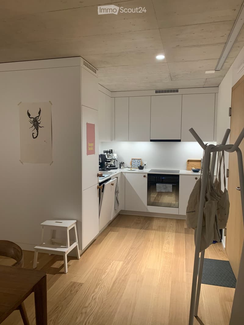 Modern kitchen with white cabinets, wooden flooring, and various appliances including a coffee maker, oven, and fridge. A small white stool and a wooden table are also visible.