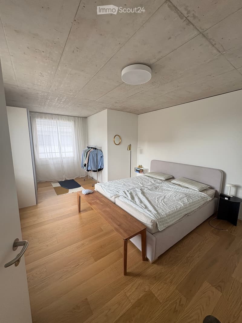 Modern bedroom with wooden flooring, a large bed with a headboard, a nightstand, a small table, a clothing rack, and a large window with sheer curtains.