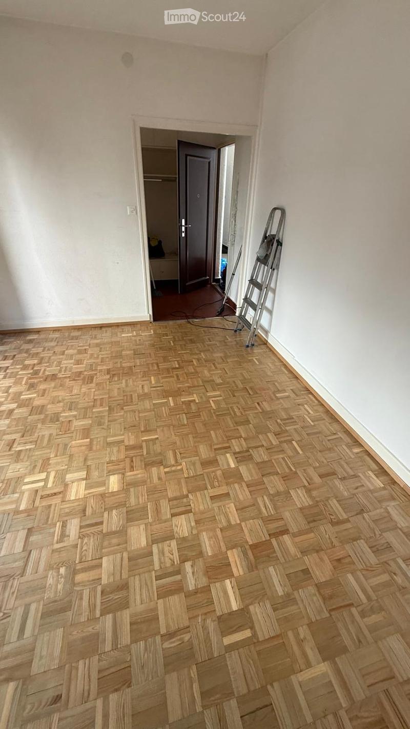 Empty room with wooden herringbone flooring, white walls, and a doorway leading to another room. A ladder is positioned against the wall near the doorway.