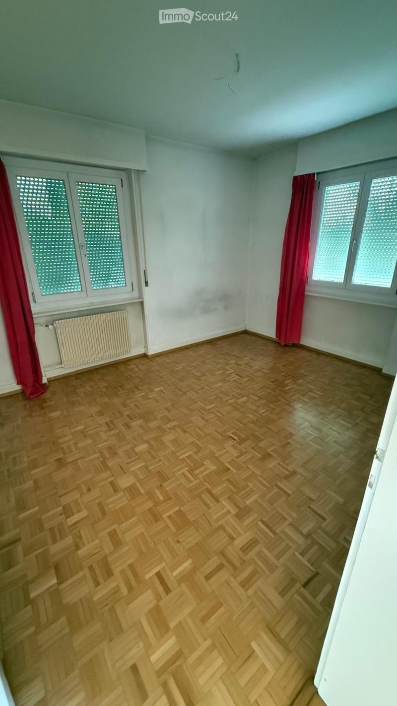 Empty room with wooden herringbone flooring, white walls, and large windows with red curtains. Radiator visible under one window.