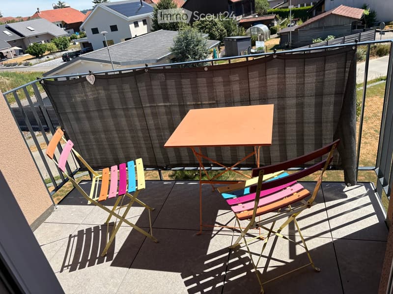 Balcony with a table and colorful folding chairs, surrounded by a metal railing and a privacy screen, overlooking a residential area.