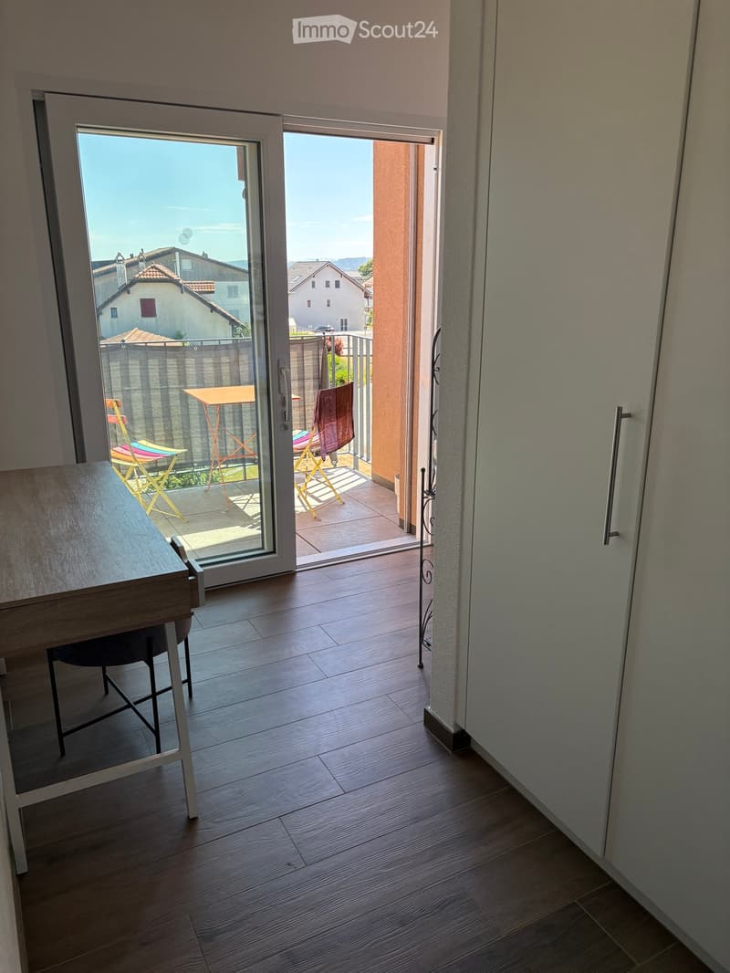 A modern interior with wooden flooring, a large sliding glass door leading to a balcony with outdoor furniture, and a small table and chair inside.