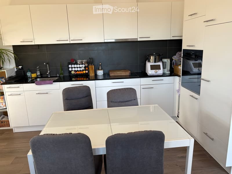 Modern kitchen with white cabinets, stainless steel appliances, a white dining table with four grey chairs, and various kitchen items on the counter.