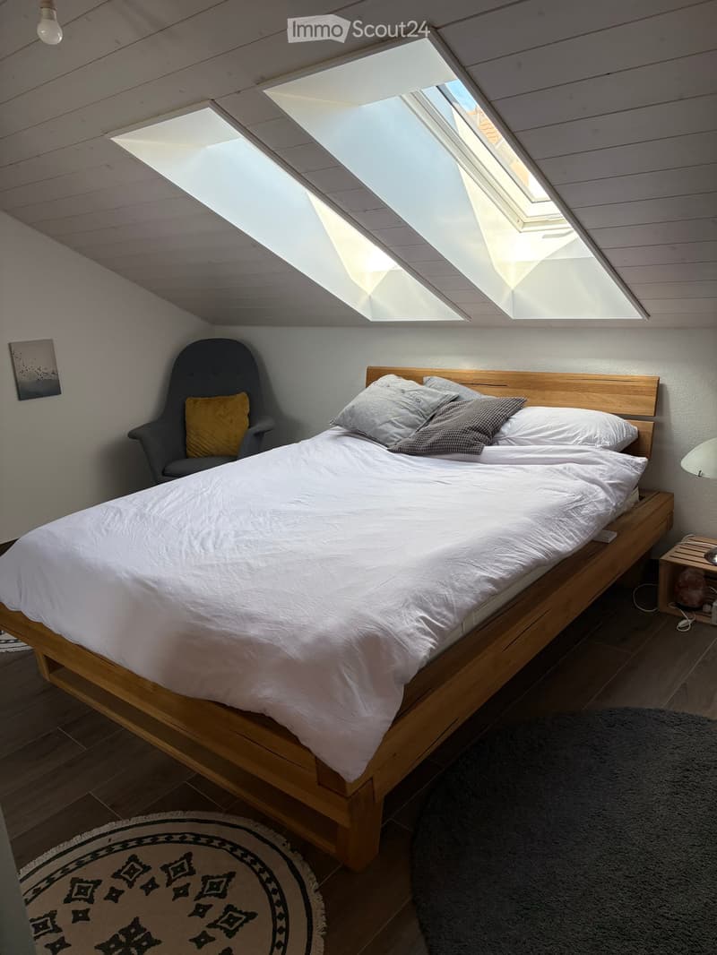 Cozy attic bedroom with wooden bed, grey armchair, nightstand, and skylight windows.