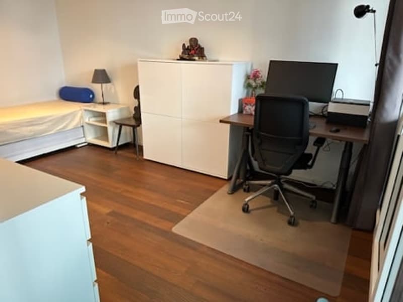 A modern, well-lit room with a wooden floor, a desk with a computer and chair, a bed with a nightstand, and a white cabinet.