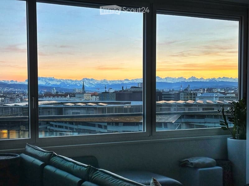 A modern balcony with large windows offering a panoramic view of a cityscape and mountains at sunset. A green sofa and a potted plant are visible.