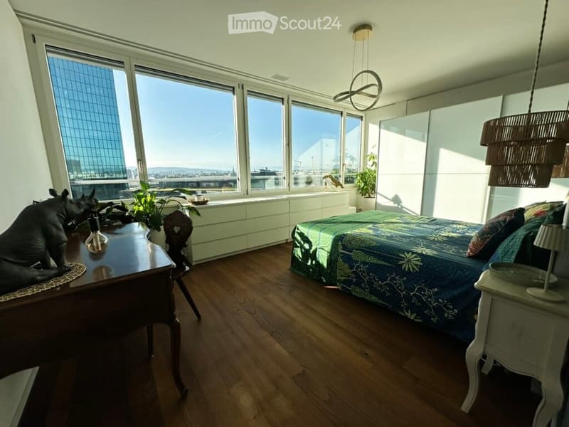 Modern bedroom with large windows offering a city view, featuring a bed with a patterned duvet, a nightstand with a lamp, a wooden table with a chair, and decorative plants.