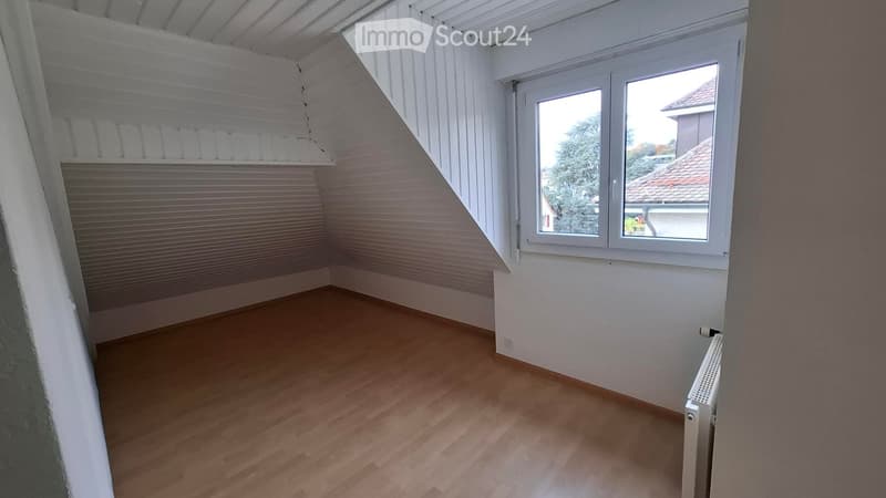 Empty attic room with wooden flooring, white walls, and a large window offering a view of the outside. The room features exposed wooden beams on the ceiling and a radiator on the wall.