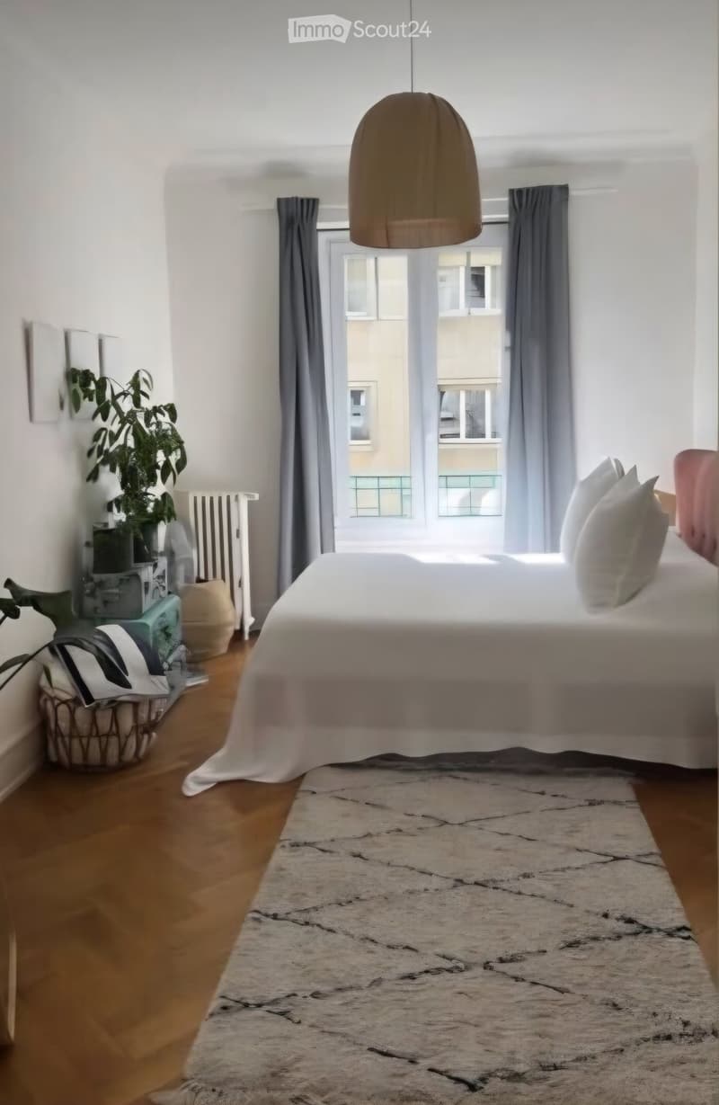 Modern bedroom with a large bed, pendant light, large window with curtains, plant, radiator, and a basket on the floor.