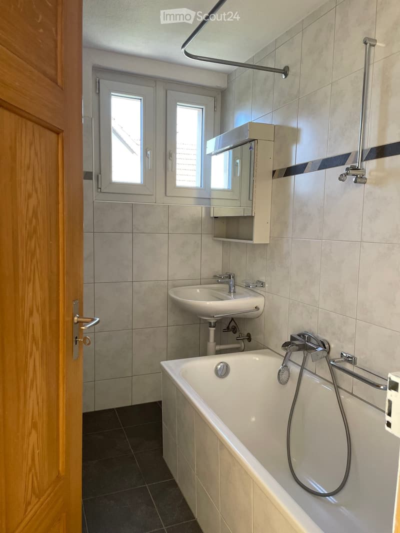 Modern bathroom with a bathtub, sink, mirror, and tiled walls. Features a wooden door and two small windows.