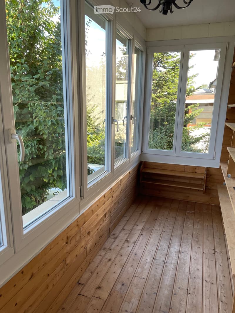Bright, wooden-floored balcony with large windows and sliding doors, offering views of greenery and neighboring buildings.