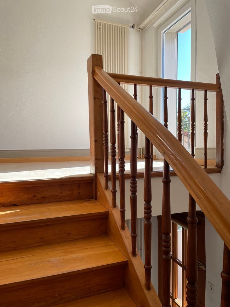 Wooden staircase with a polished handrail and balusters, leading to an upper level. The walls are painted white, and a window at the top of the stairs allows natural light to enter.